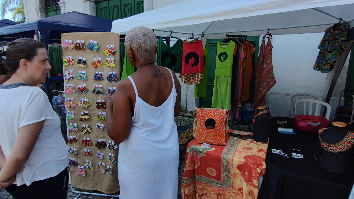 Mulheres em uma feira de artesanato observam uma banca que vende brincos coloridos e roupas vibrantes de estampas étnicas