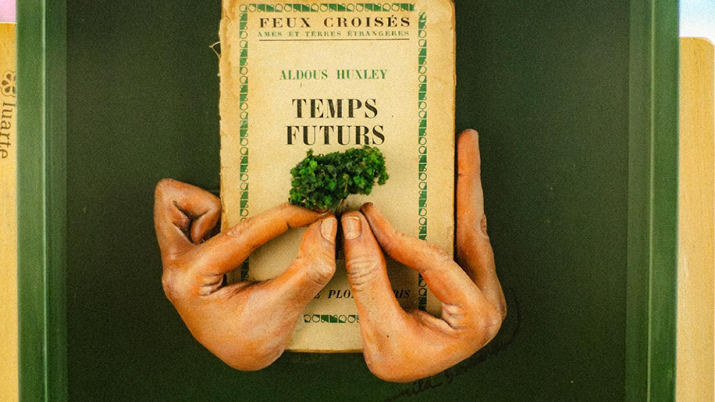Uma fotografia em close-up de uma obra de arte em uma moldura verde e preta, onde um livro antigo intitulado "TEMPS FUTURS" (Tempos Futuros) é segurado por mãos esculpidas que pinçam uma pequena folhagem, no centro