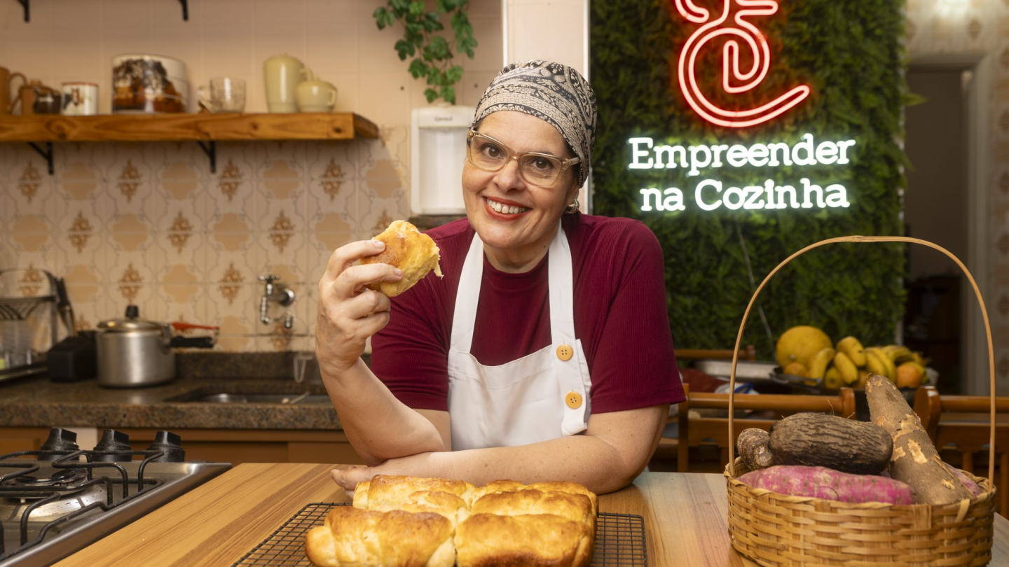A imagem mostra a empreendedora Ana Cláudia Siqueira em uma cozinha, sorrindo e segurando um pedaço do famoso Pão de Cara de Santos, com pães recém-assados em uma grade à frente