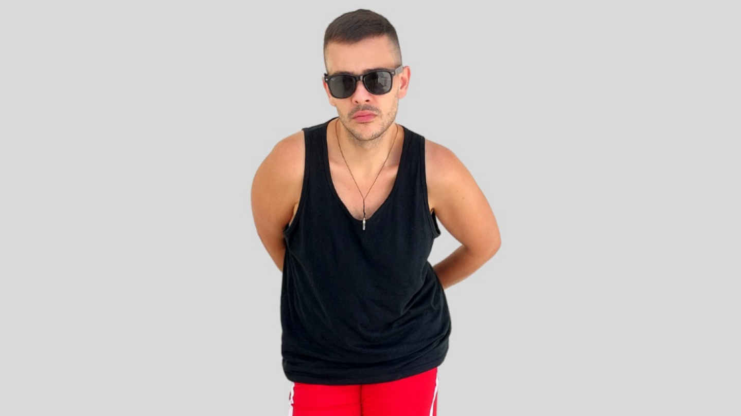 A imagem mostra um homem em pé diante de um fundo cinza claro, usando óculos escuros, regata preta e shorts vermelhos, com as mãos para trás e expressão séria