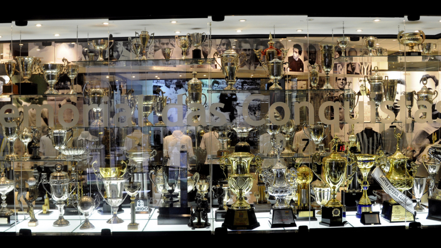 Vitrine ampla repleta de troféus de diferentes tamanhos e formatos, todos brilhantes e organizados em prateleiras de vidro. Ao fundo, fotos antigas em preto e branco compõem o cenário do memorial esportivo