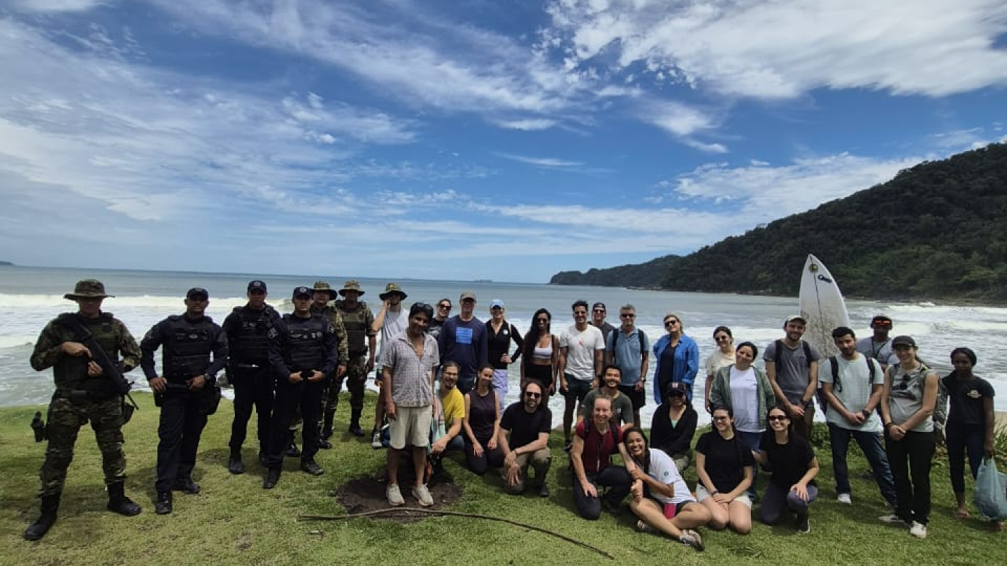 Um grupo grande de pessoas posa na grama, com militares e civis, em frente a uma paisagem de praia e montanha.