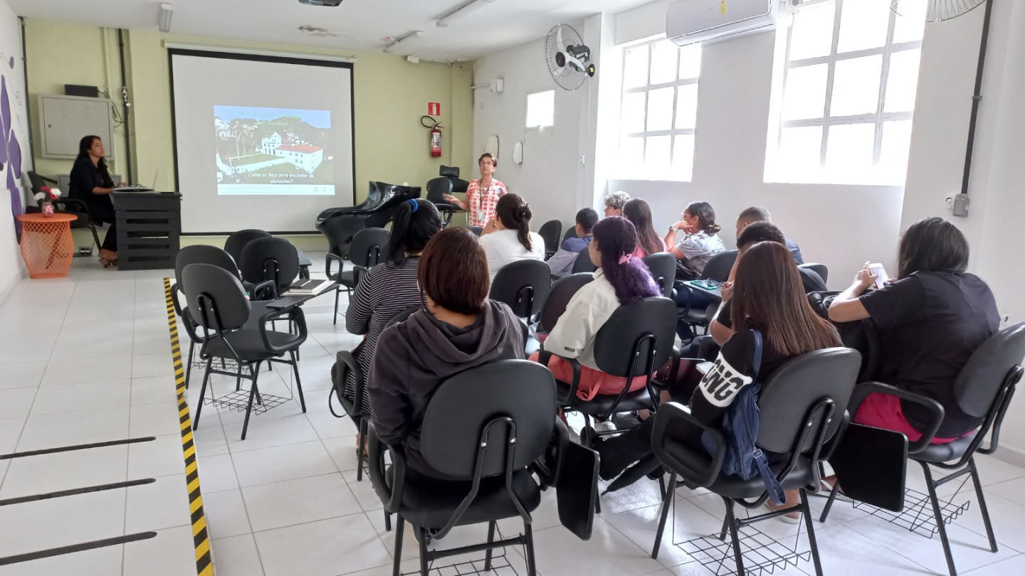 Imagem mostra uma mulher em pé ministrando uma palestra para um grupo de pessoas sentadas em uma sala de aula. Ao fundo, há uma tela de projeção com uma apresentação sobre atendimento a visitantes.