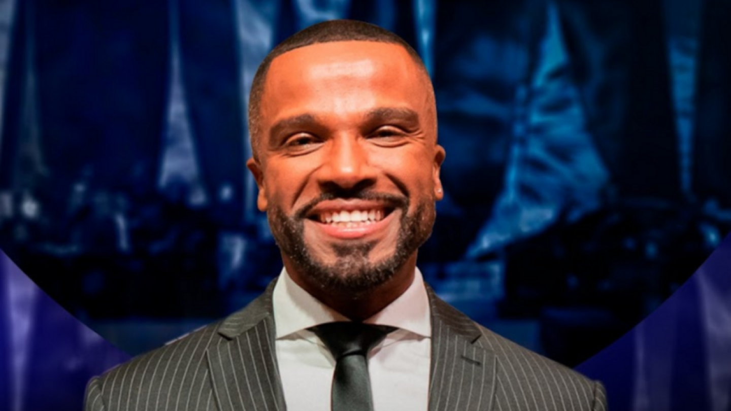 Homem sorridente de terno e gravata diante de um fundo escuro com iluminação de palco