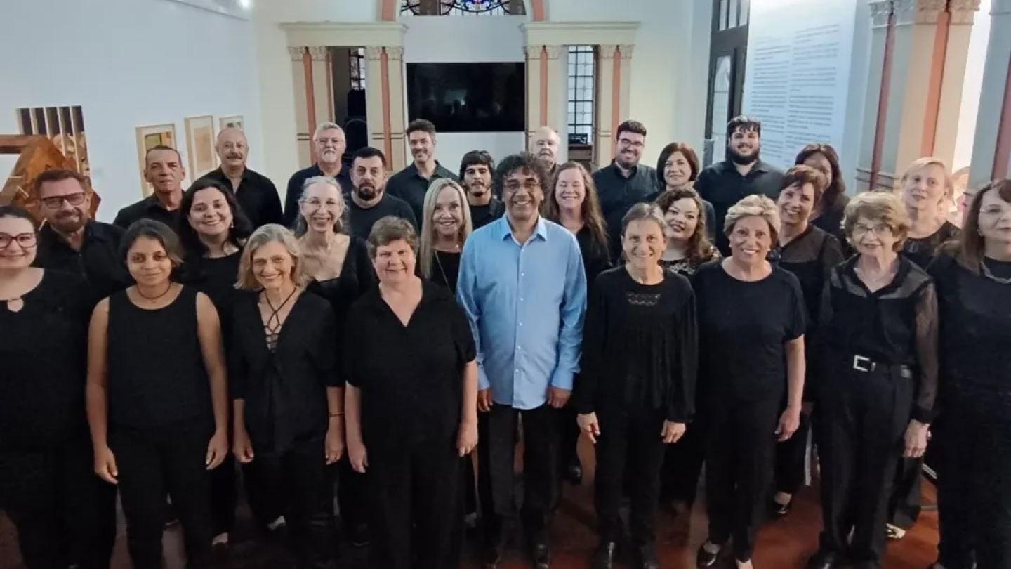 Foto de um grupo de pessoas de um coral, vestindo preto, sorrindo e posando em um salão ornamentado
