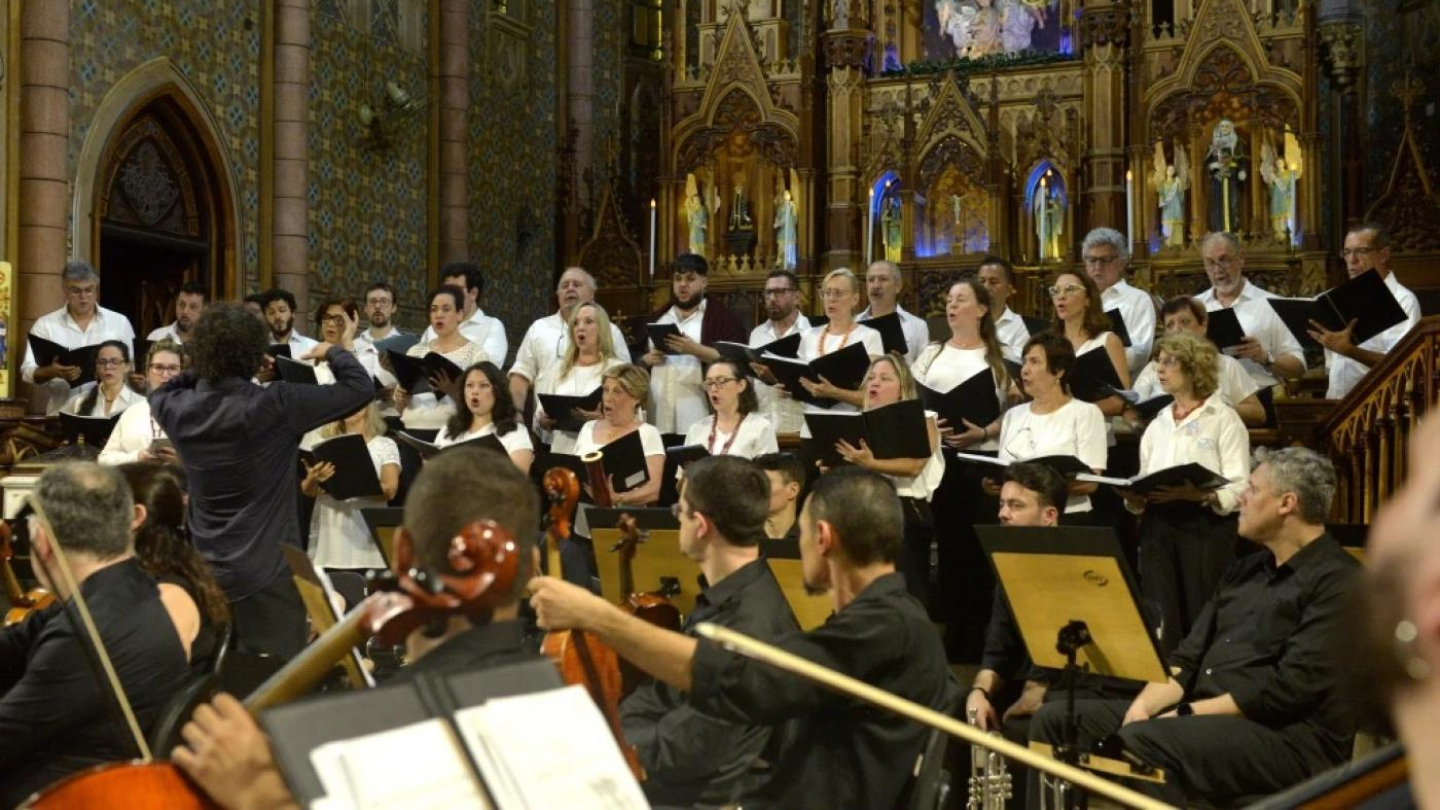 Um coral e uma orquestra se apresentam em um ambiente interno de uma igreja ricamente decorada