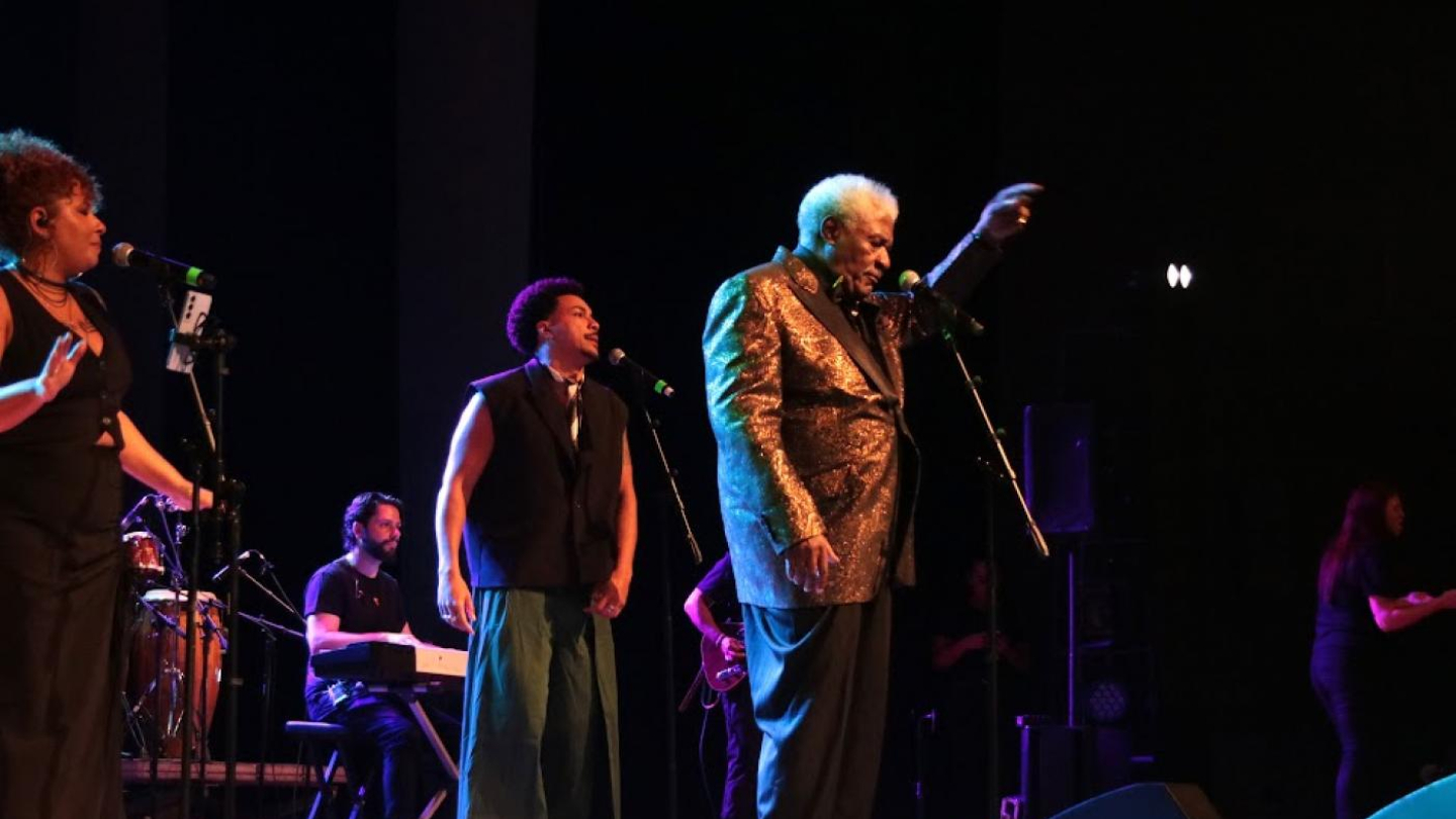 Um cantor idoso, com cabelos grisalhos e um paletó de lantejoulas douradas, se apresenta em um palco com um microfone na mão e apontando para o lado, ao lado de outros músicos