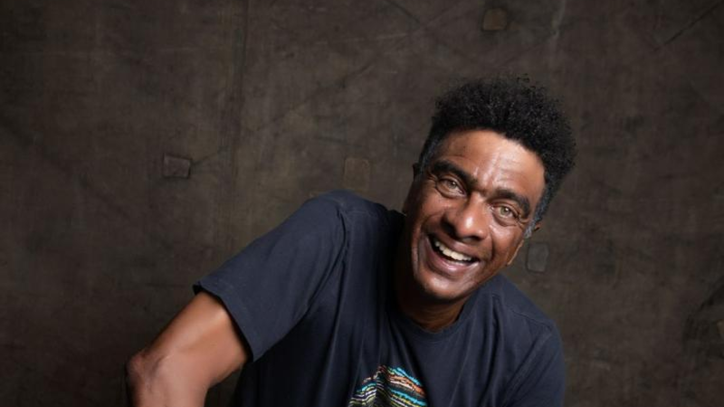 Hélio de la Peña, ator e humorista negro de cabelos curtos encaracolados, está sentado em um banquinho de madeira clara. Ele veste uma camiseta preta com uma estampa colorida e calças jeans. Ele sorri amplamente e olha diretamente para a câmera, com o corpo inclinado ligeiramente para frente. O fundo é uma parede ou tecido de tom marrom escuro e texturizado