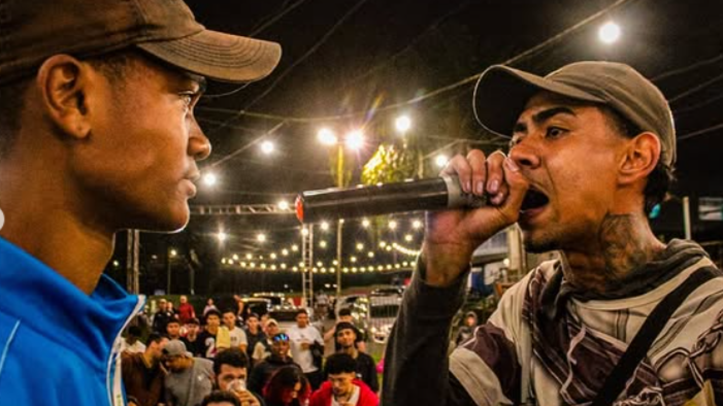 Dois homens de boné se encaram, um deles segurando um microfone, enquanto uma plateia observa