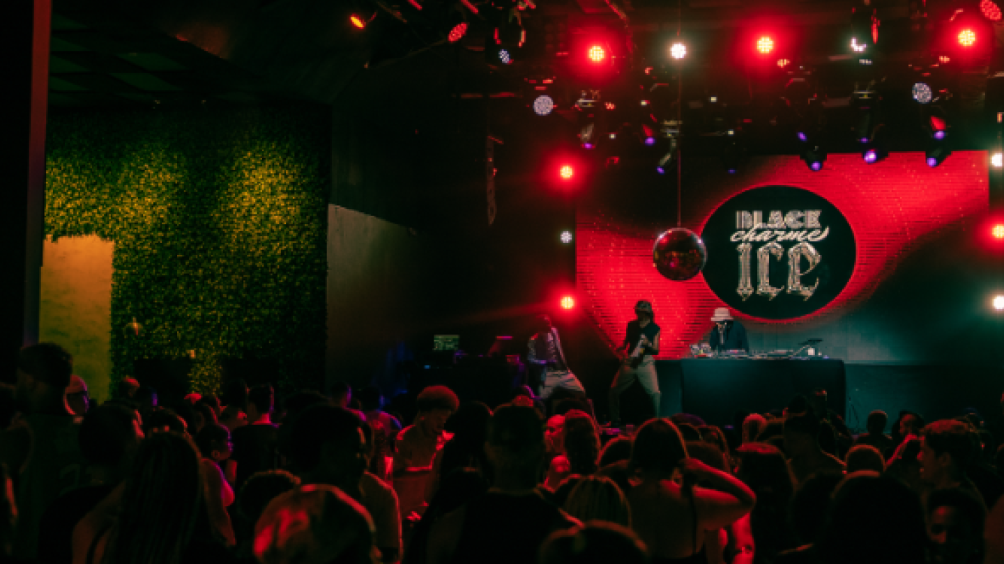A imagem de uma festa noturna mostra uma multidão dançando na frente de um palco com luzes vermelhas, uma bola de espelhos e a inscrição "BLACK CHARME ICE" no painel de LED