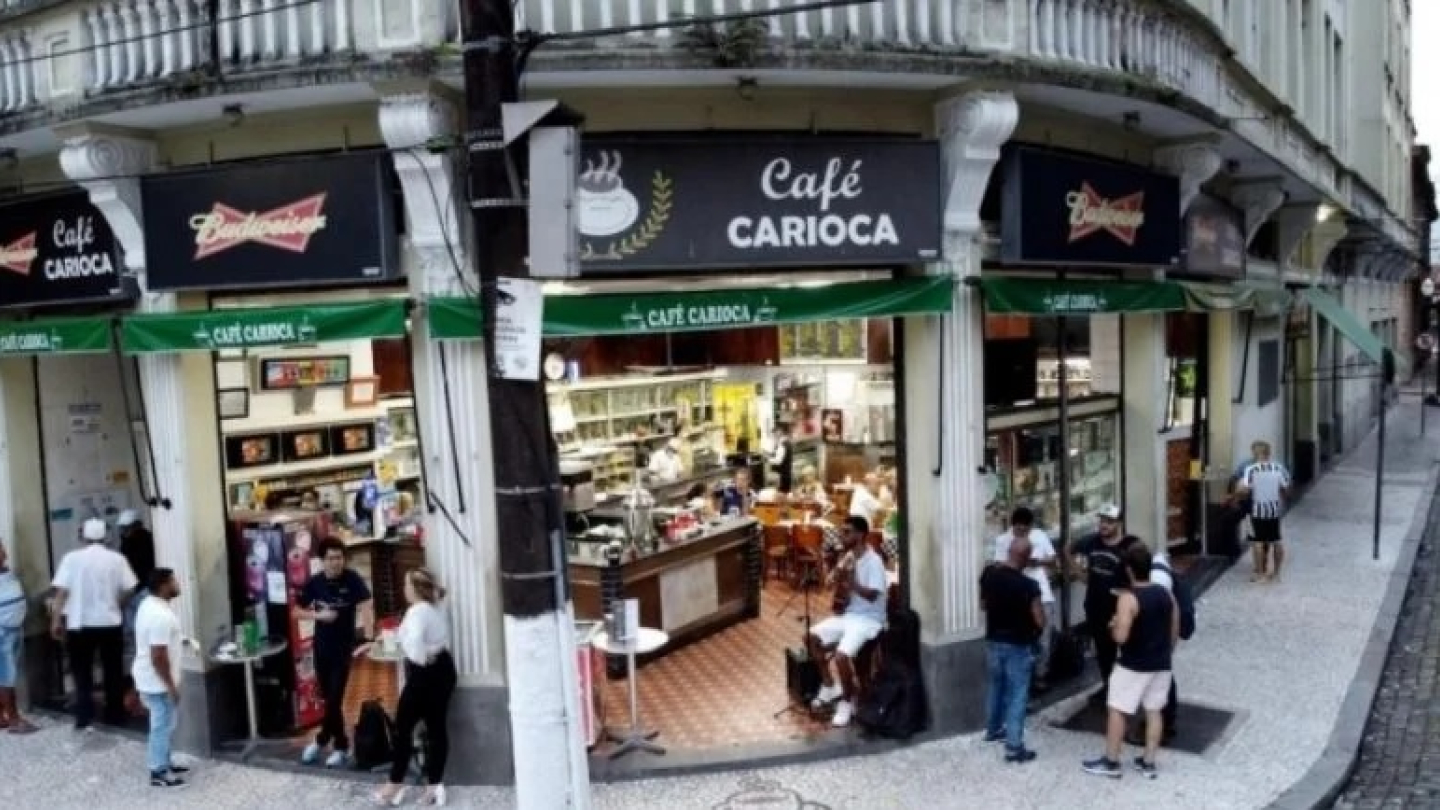Uma foto de ângulo aberto mostra a fachada de esquina do tradicional "Café Carioca", onde pessoas estão socializando do lado de fora e no interior, e há um músico tocando violão na entrada do estabelecimento