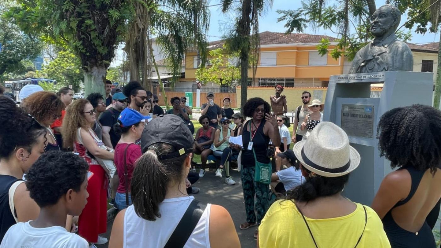 A imagem mostra um grupo de pessoas reunido ao ar livre, em um parque ou praça ensolarada, participando de um tour guiado, ao lado de busto de um homem em um pedestal de pedra, num ambiente arborizado