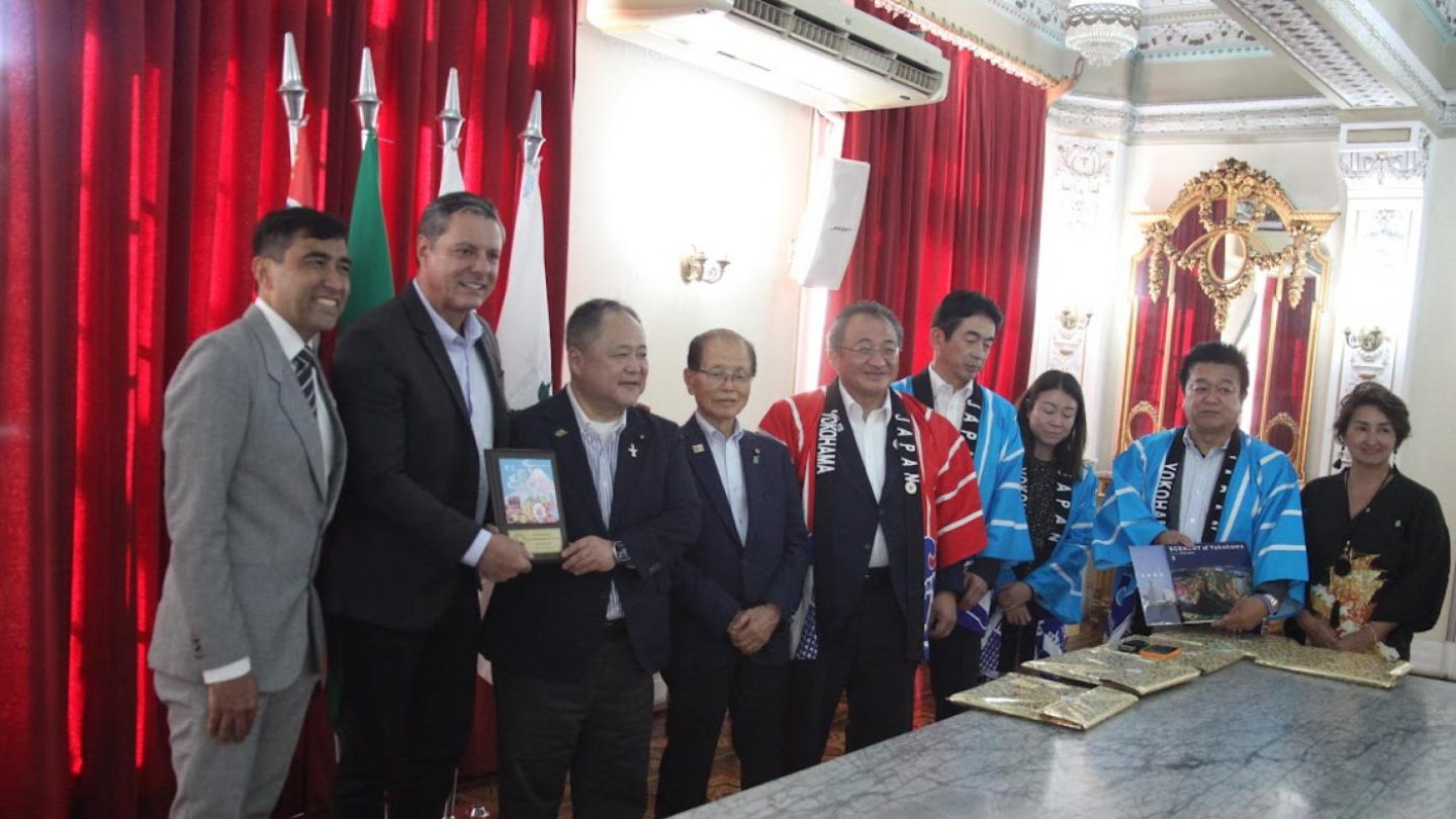 Um grupo de cerca de dez pessoas, algumas de terno e outras vestindo faixas e casacos happi japoneses coloridos, estão em uma sala formal com cortinas vermelhas e decoração ornamentada, posando em torno de uma mesa de mármore