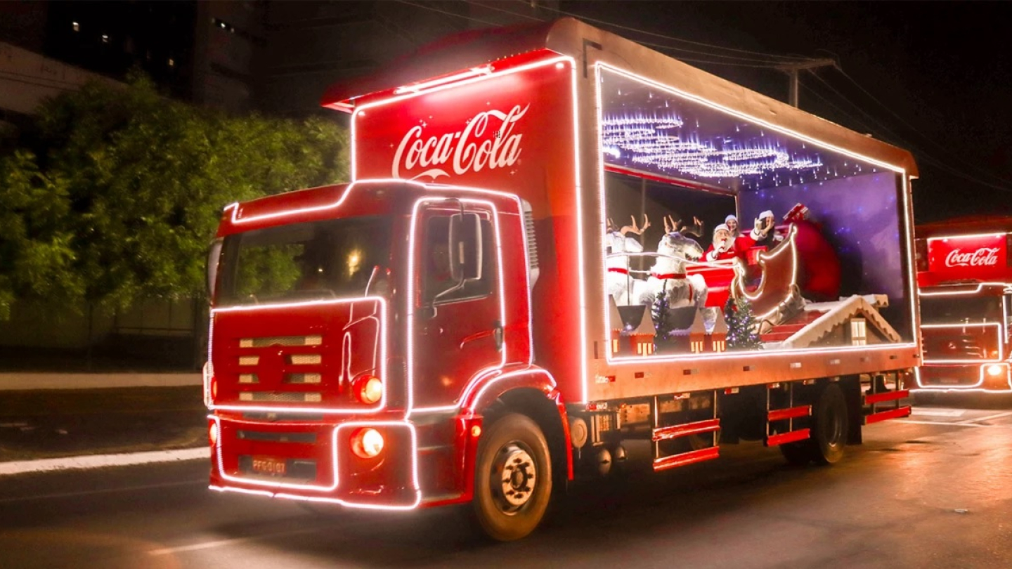 Dois caminhões vermelhos da Coca-Cola estão iluminados com luzes de LED brancas contornando a cabine e a carroceria, sendo que o primeiro transporta em sua parte traseira aberta uma cena natalina com a representação do Papai Noel em um trenó