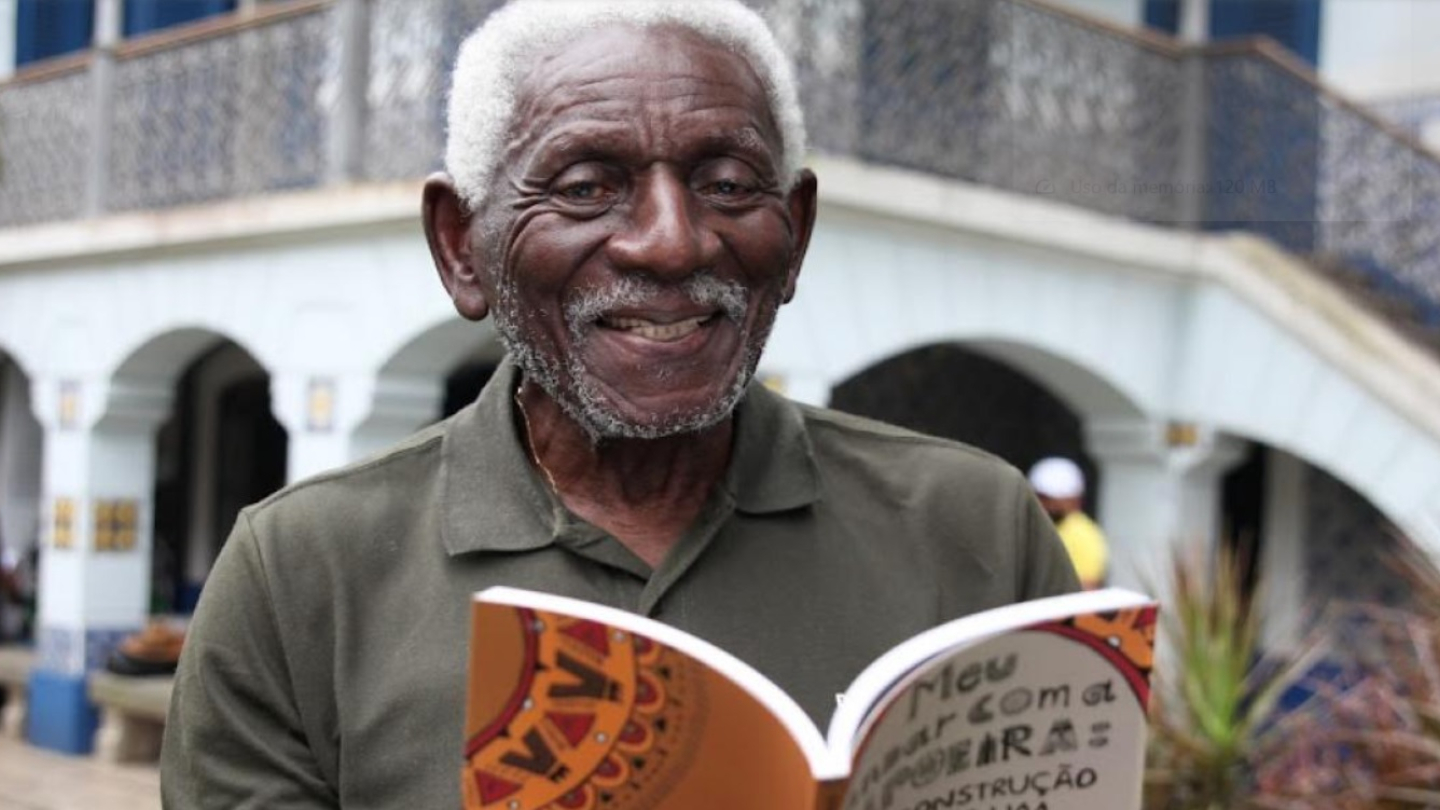 Um senhor negro de cabelos grisalhos sorri enquanto segura um livro aberto em frente a uma casa com azulejos azuis e brancos