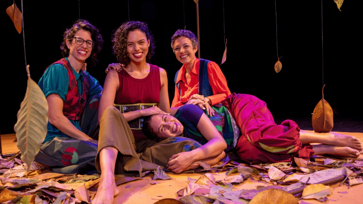 Quatro atrizes sorridentes, descalças e em figurinos coloridos, posam em um palco coberto por folhas secas penduradas e espalhadas, em um cenário que sugere uma peça teatral