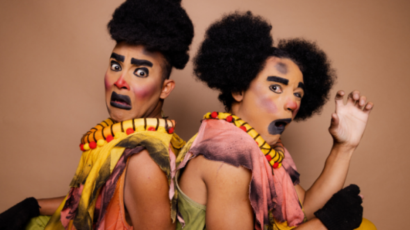 Dois artistas de teatro estão lado a lado e de costas um para o outro, ambos com maquiagem expressiva, perucas crespas e vestimentas coloridas e rasgadas, olhando para a câmera com expressões de espanto