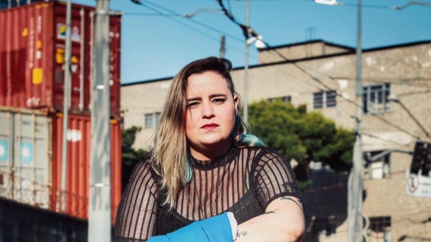 Uma mulher com cabelo loiro e mechas coloridas, vestindo uma blusa preta transparente e calças pretas, senta-se em um bloco de concreto segurando um paletó azul em uma área urbana com contêineres e edifícios ao fundo