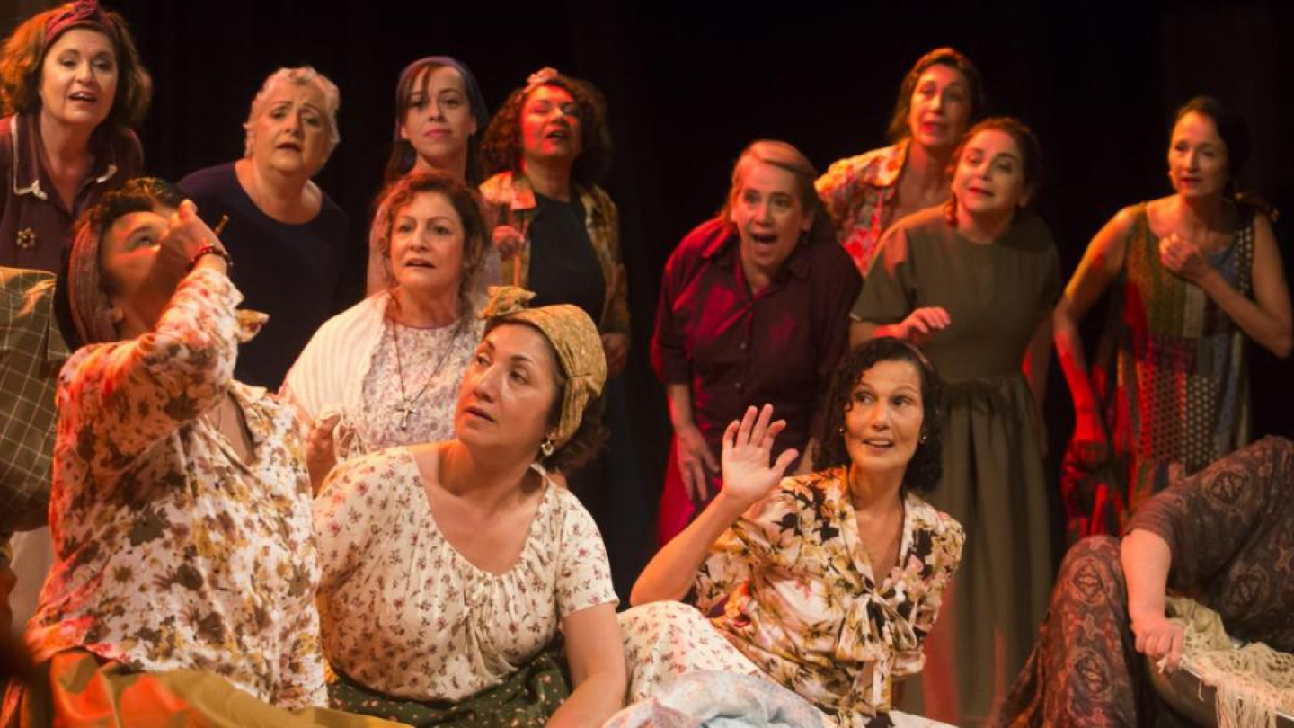 Um grupo de cerca de doze mulheres, vestidas com trajes de época (incluindo lenços na cabeça e vestidos estampados) e com expressões dramáticas, posicionado num palco com fundo escuro