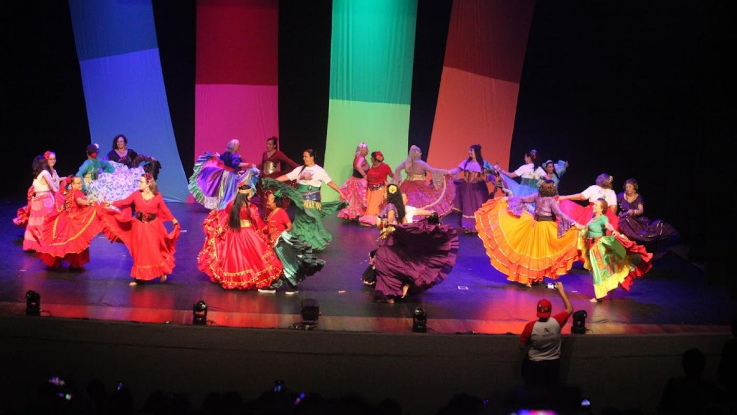 Um grande grupo de dançarinos em trajes folclóricos coloridos e saias rodadas se apresenta em um palco, iluminado por luzes de fundo em tons de vermelho, verde e azul