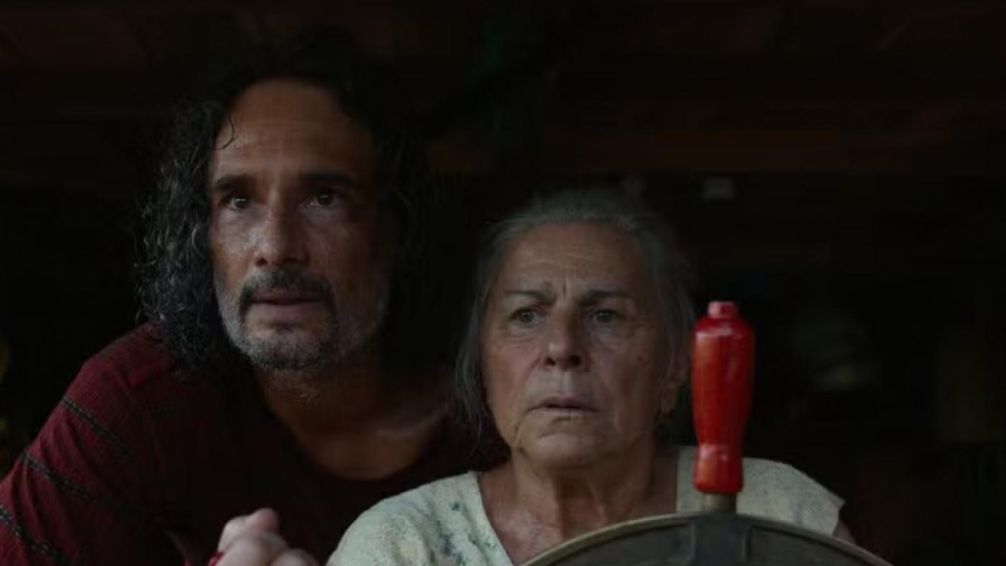 Um homem de cabelos longos e escuros e uma mulher idosa de cabelos grisalhos olham fixamente para a frente, com a mulher segurando o timão