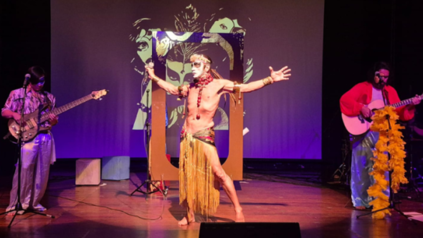 Cena de apresentação musical teatralizada, com três artistas no palco — um ao centro em performance expressiva e dois tocando instrumentos, sob iluminação colorida e cenário artístico ao fundo