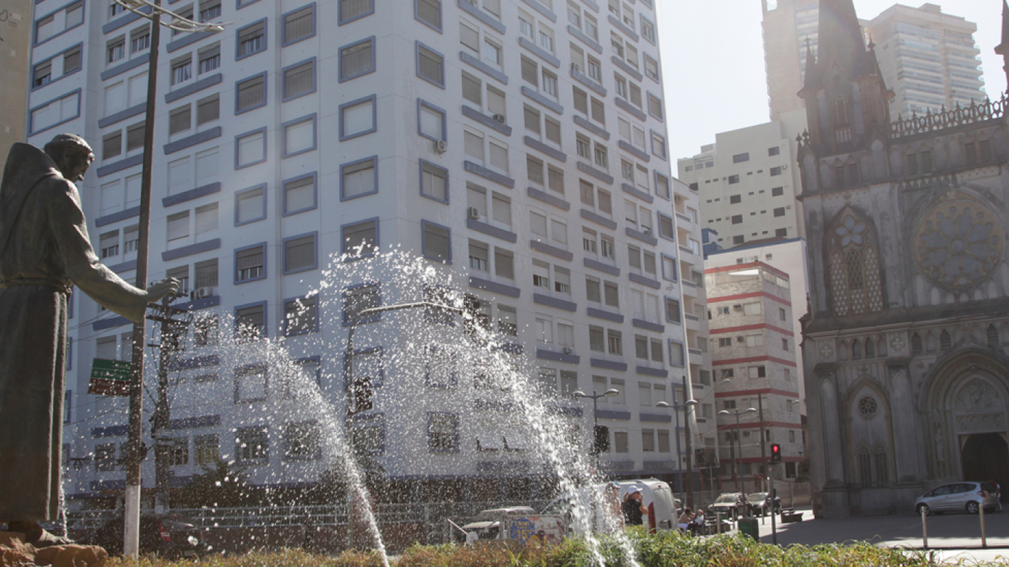 Estátua de frade em primeiro plano, com jatos d'água de uma fonte, em frente à fachada neogótica da Basílica de Santo Antônio do Embaré e prédios modernos. O céu claro e a luz forte destacam o contraste da arquitetura histórica e contemporânea na orla de Santos.