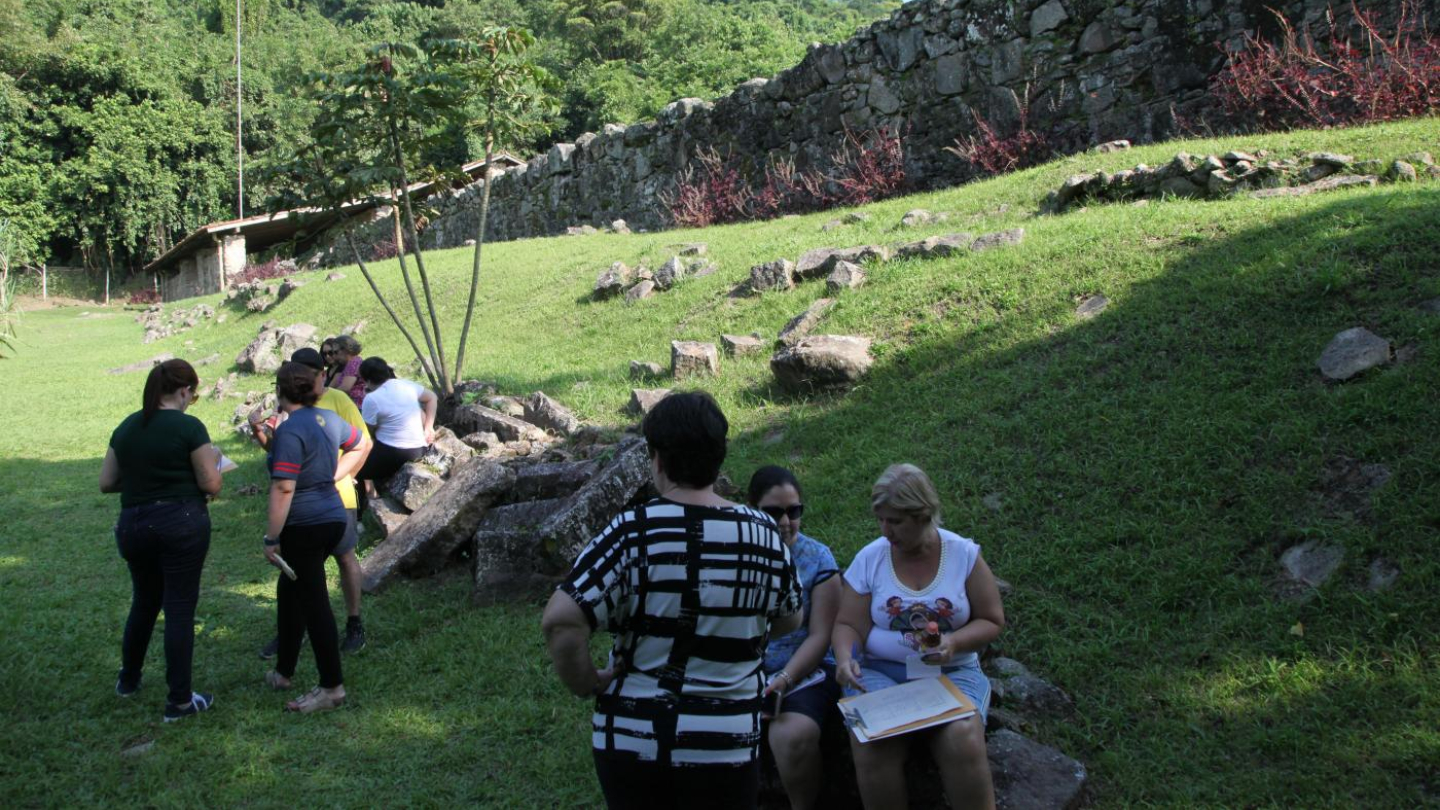  Uma fotografia mostra um grupo de pessoas reunidas em um campo gramado, algumas conversando e outras sentadas em pedras, com uma antiga muralha de pedra e uma encosta de montanha densamente arborizada ao fundo sob um céu azul