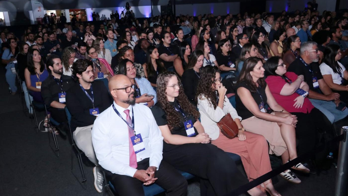 Uma grande plateia de pessoas, incluindo o primeiro plano com crachás, está sentada atenta em cadeiras em um auditório escuro durante um evento ou palestra