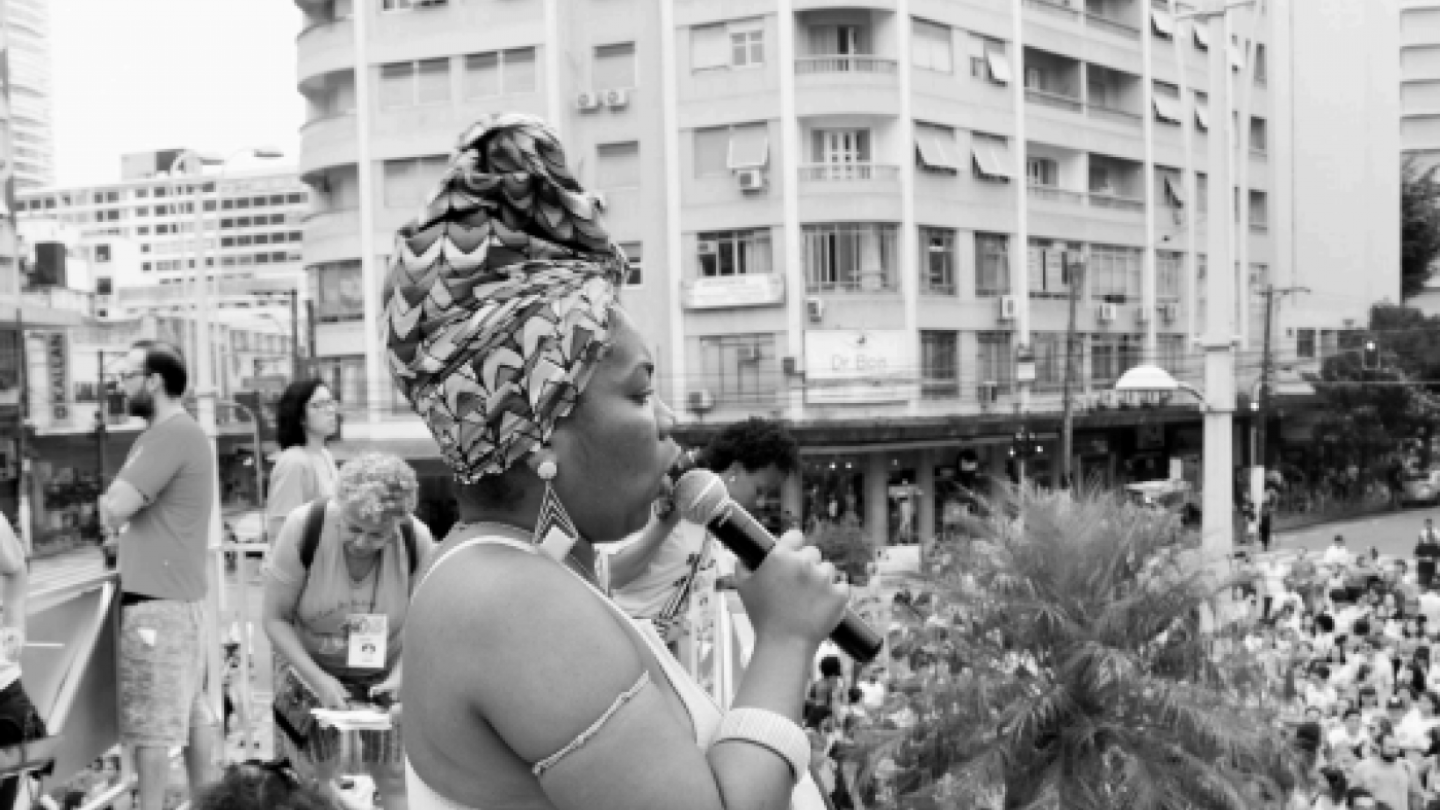 Em uma foto em preto e branco, uma mulher negra com um turbante estampado e brincos de argola canta em um microfone enquanto está em um palco, com uma multidão e prédios urbanos no fundo
