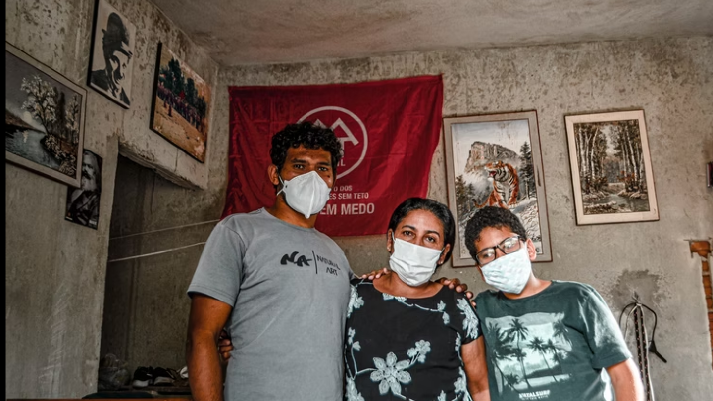 Uma família (homem, mulher e adolescente), todos usando máscaras de proteção, posa em frente a uma bandeira vermelha do MTST (Movimento dos Trabalhadores Sem Teto) em um interior com quadros e paredes rústicas