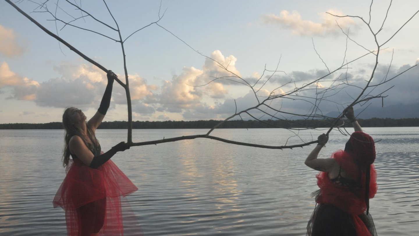 Duas mulheres vestidas de vermelho e preto segurando galhos secos sobre um corpo d'água ao pôr do sol