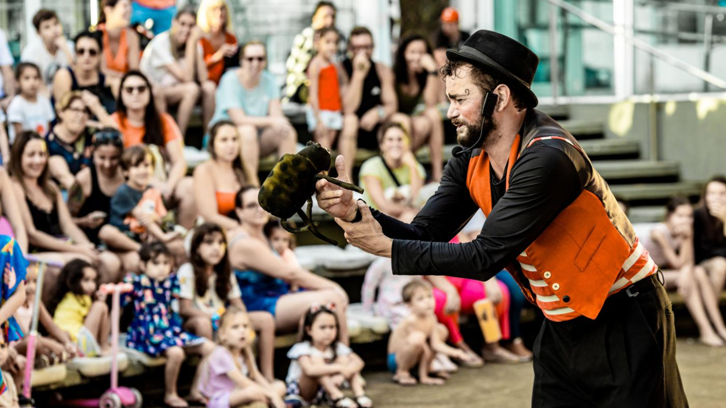 Um artista de rua, vestindo colete laranja e chapéu preto, está se apresentando para uma grande plateia de adultos e crianças sentadas em degraus ao ar livre