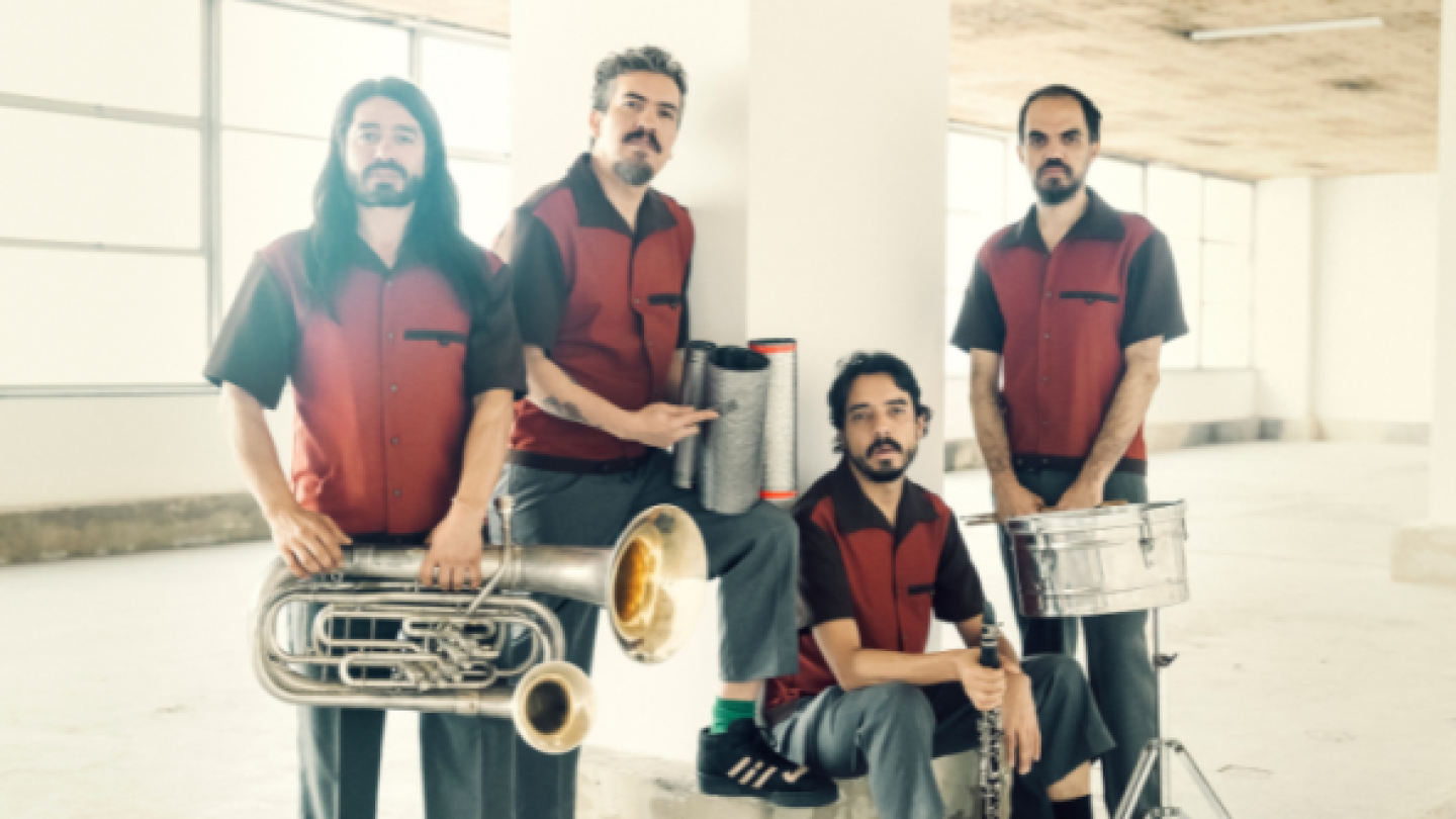 Quatro músicos barbados, todos vestindo camisas de botão vermelhas e pretas, posam segurando instrumentos musicais