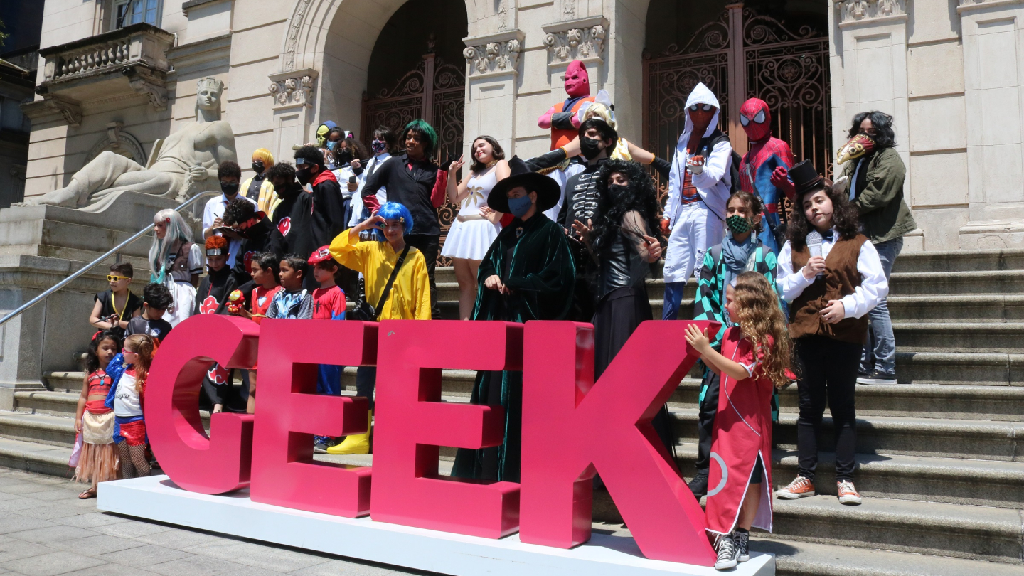 Diversas pessoas vestindo fantasias de cosplay de personagens de cultura pop reunido nas escadarias de um edifício histórico e, em primeiro plano, há um grande letreiro rosa vibrante com a palavra "GEEK"