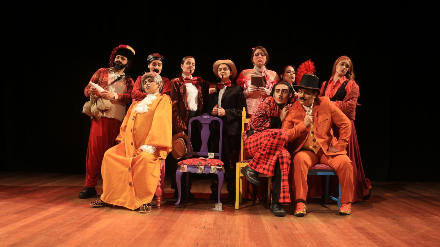 A imagem captura um grupo de dez atores de teatro, alguns sentados em cadeiras de cores e estilos variados, vestidos com figurinos coloridos e extravagantes de época, com uma predominância de tons quentes como vermelho e laranja, em um palco de madeira com fundo preto