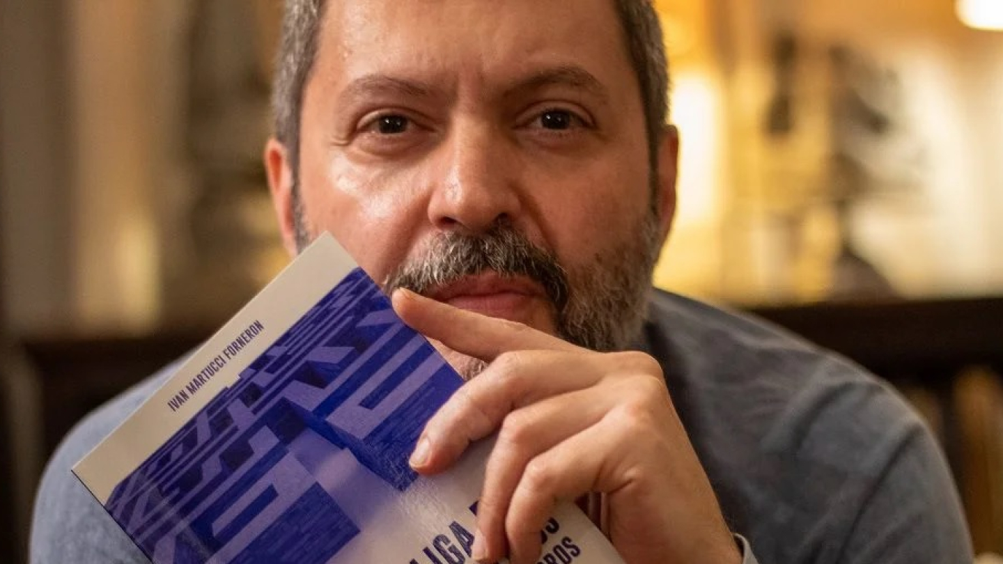 Um homem barbudo de meia-idade segura um livro, cobrindo parte do rosto, e olha diretamente para a câmera