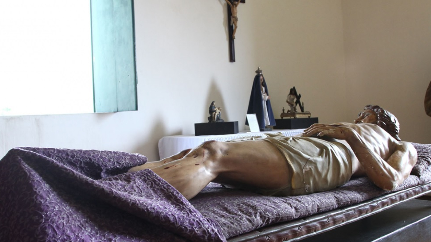 Uma cena de arte sacra em um quarto iluminado pela luz de uma janela aberta, onde uma escultura de Jesus morto repousa em um leito coberto com um tecido roxo, com outras estátuas e um crucifixo na parede ao fundo