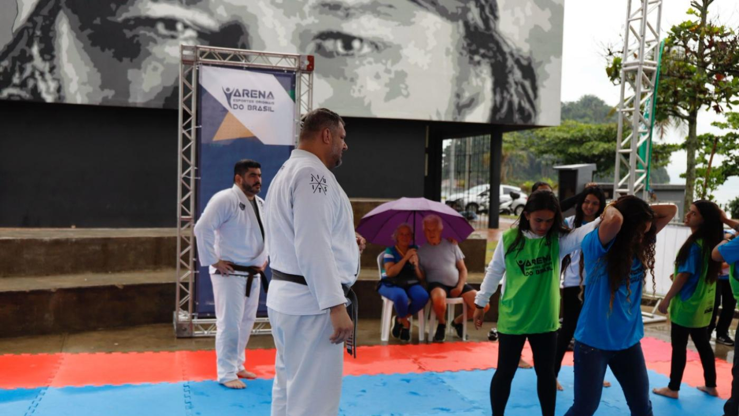 Pessoas praticando Jiu-Jitsu sobre um tatame ao ar livre, com um grande mural em preto e branco ao fundo