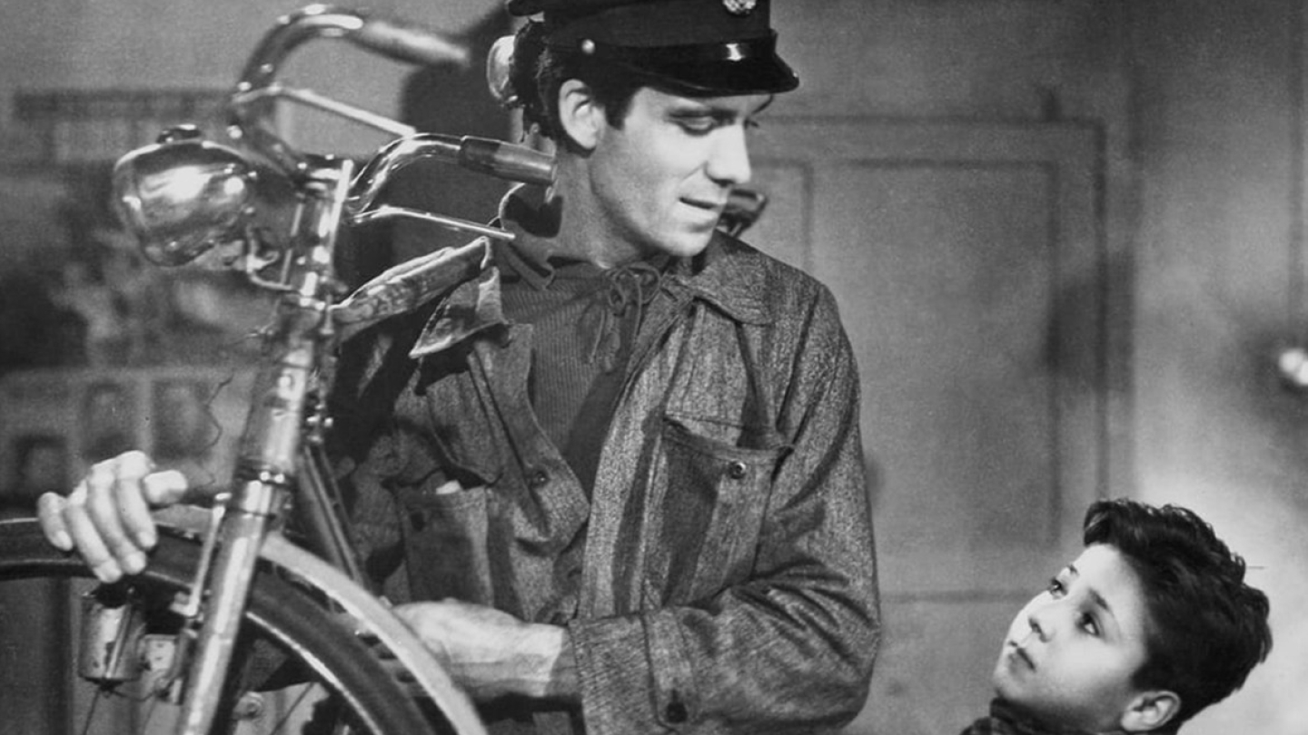 Imagem em preto e branco do filme Ladrões de Bicicletas mostra Antonio Ricci com seu uniforme de trabalho, segurando a bicicleta roubada, e olhando com ternura para seu filho Bruno