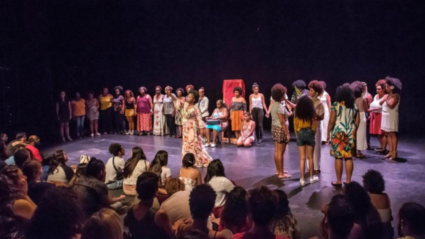 Um grande grupo de mulheres negras, algumas de mãos dadas, estão reunidas em um palco circular sob iluminação central em um teatro de fundo escuro, com uma plateia sentada no chão em primeiro plano