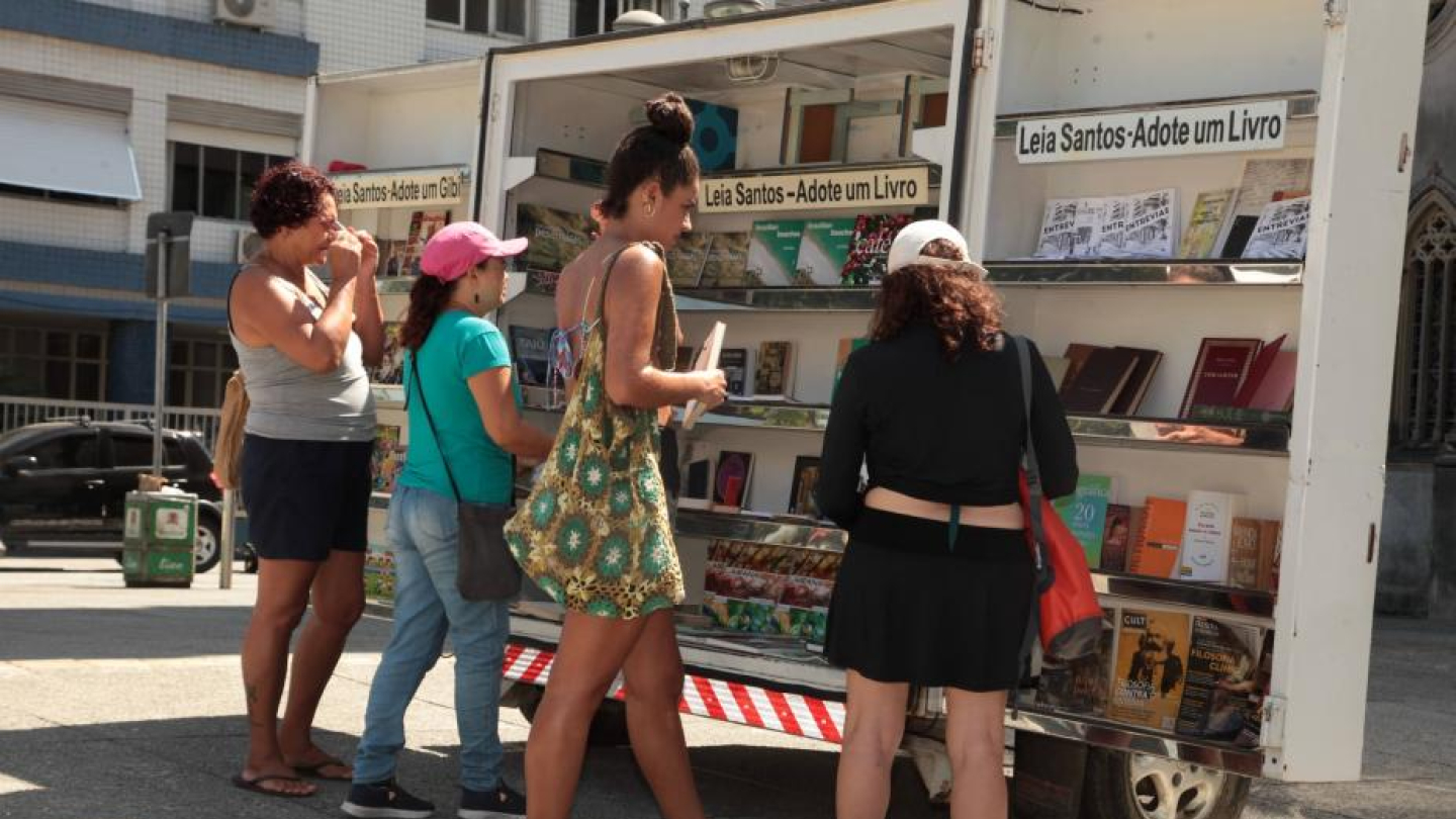 Quatro mulheres estão em uma rua ensolarada examinando livros em um veículo adaptado como estante de livros, com as inscrições "Leia Santos – Adote um Livro"