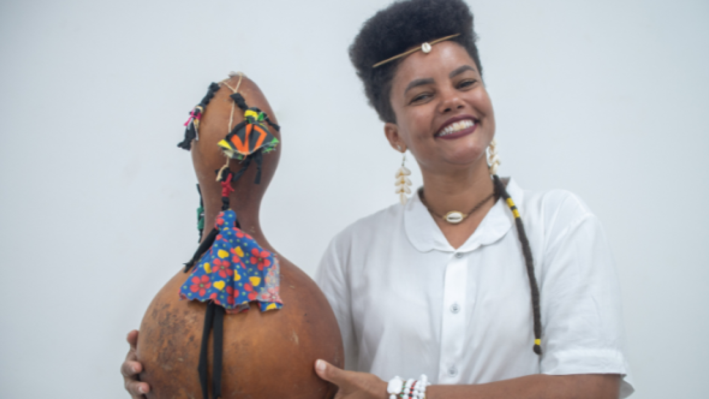 Uma mulher sorridente com cabelo afro, acessórios tradicionais e uma camisa branca segura uma cabaça decorada em frente a um fundo branco