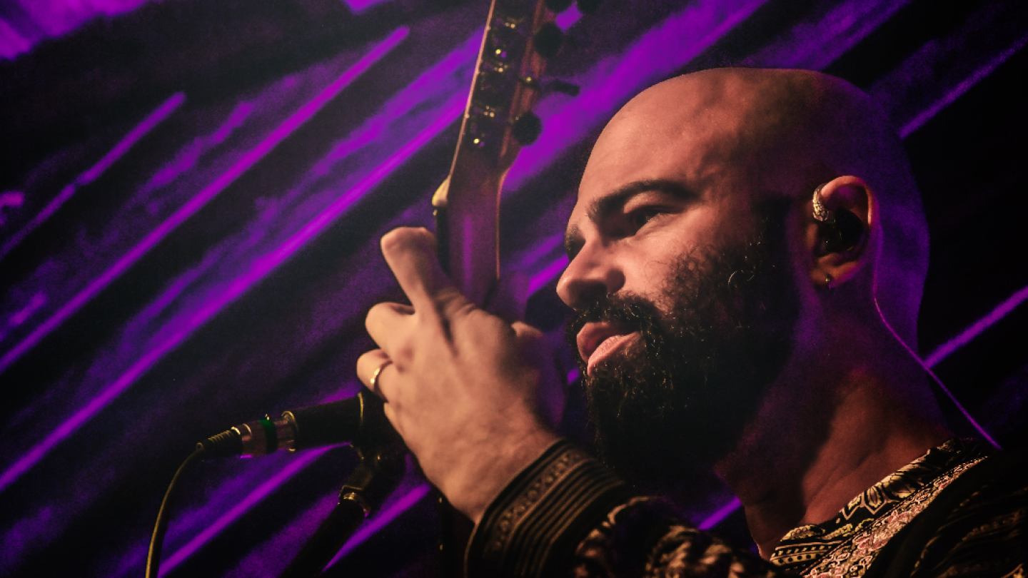 Um músico barbudo, de cabeça raspada, toca uma guitarra em um palco, iluminado por luzes roxas que criam um efeito de fundo listrado