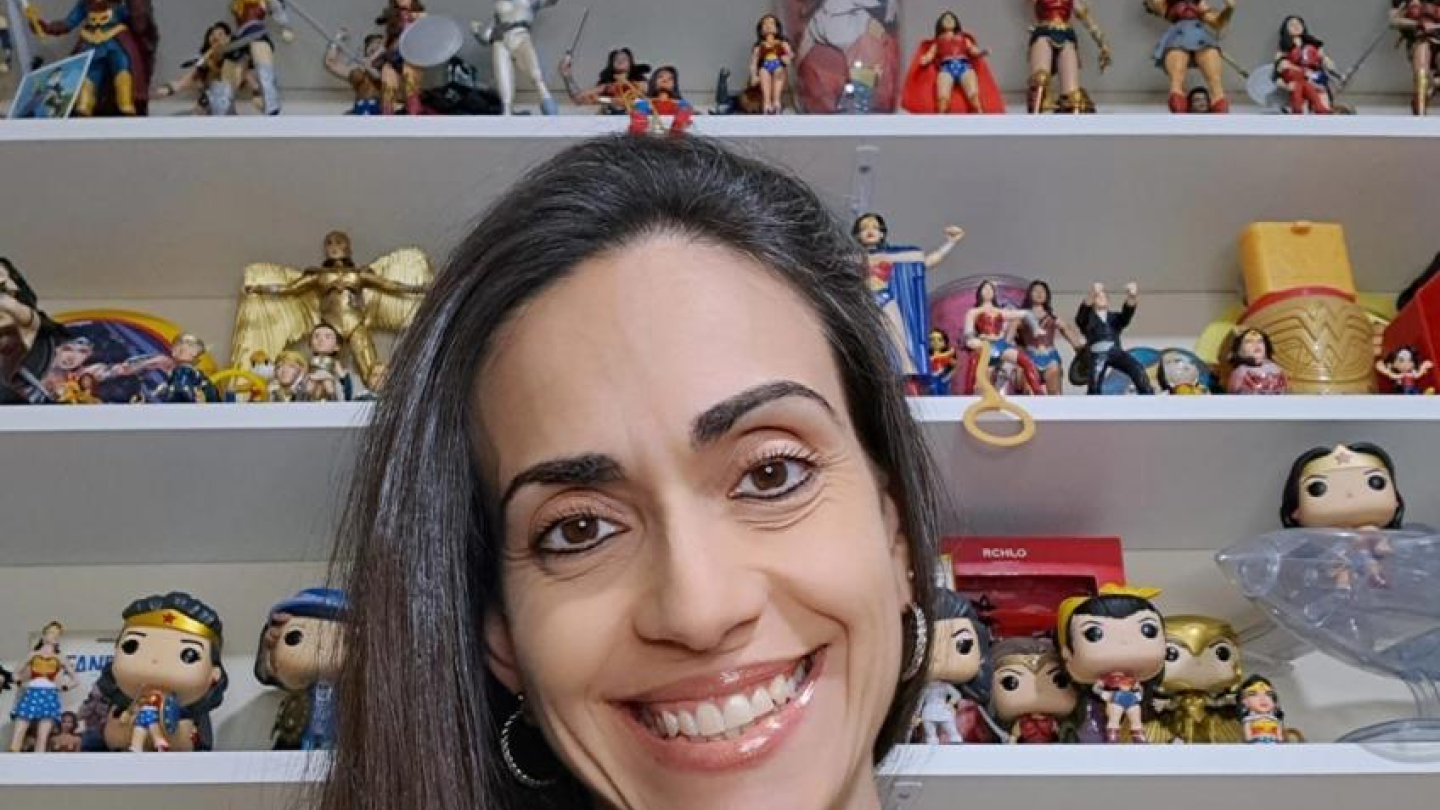 mulher sorridente em primeiro plano, tirando uma selfie em frente a uma prateleira repleta de colecionáveis da Mulher-Maravilha