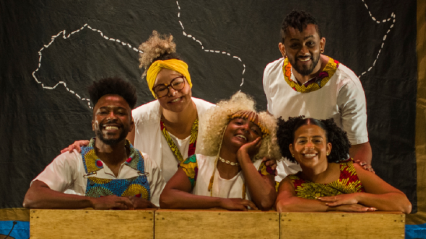 Cinco artistas sorridentes, vestindo roupas e acessórios de estampa afro, posam juntos em um palco com um mapa da África desenhado em giz no fundo preto