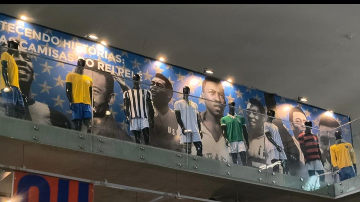 Uma foto de interior em ângulo inferior mostra uma exposição suspensa no Museu Pelé, apresentando manequins vestindo réplicas de diversas camisas usadas pelo jogador sobre um painel azul decorado com estrelas e fotos em preto e branco de Pelé, e o título "TECENDO HISTÓRIAS: AS CAMISAS DO REI PELÉ"