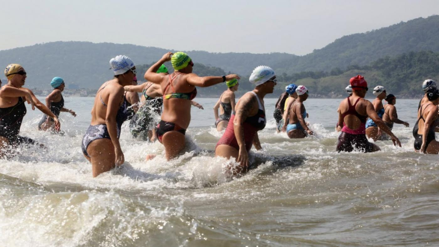 Grupo de mulheres nadadoras entrando no mar, durante uma prova de natação em águas abertas, com um cenário de montanhas ao fundo