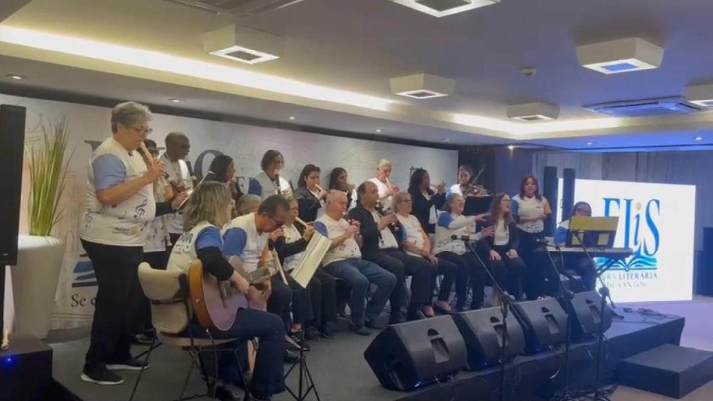 Um grupo de músicos de diferentes idades, alguns sentados e outros em pé, se apresenta com instrumentos variados (incluindo flautas e violão) em um palco com iluminação clara