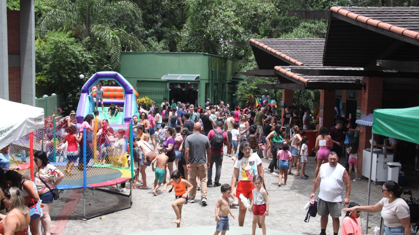 Imagem mostra uma grande área ao ar livre repleta de pessoas, incluindo muitas crianças e famílias. Há brinquedos infláveis coloridos, como um escorregador e uma cama elástica, e várias barracas montadas ao redor. O ambiente é ensolarado e cercado por árvores, transmitindo clima de diversão e lazer em família.